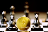 A gold Ethereum token is prominently placed on a black and white chessboard surrounded by several metallic chess pieces. The focus is on the Ethereum symbol on the coin, while the chess pieces appear slightly out of focus in the background. The lighting emphasizes the reflective surfaces of both the coin and the chess pieces.