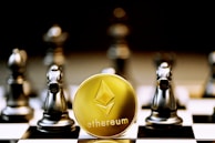 A gold Ethereum token is prominently placed on a black and white chessboard surrounded by several metallic chess pieces. The focus is on the Ethereum symbol on the coin, while the chess pieces appear slightly out of focus in the background. The lighting emphasizes the reflective surfaces of both the coin and the chess pieces.