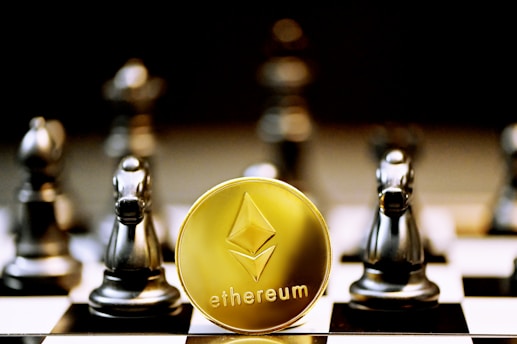 A gold Ethereum token is prominently placed on a black and white chessboard surrounded by several metallic chess pieces. The focus is on the Ethereum symbol on the coin, while the chess pieces appear slightly out of focus in the background. The lighting emphasizes the reflective surfaces of both the coin and the chess pieces.