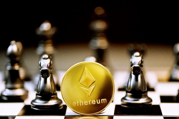 A gold Ethereum token is prominently placed on a black and white chessboard surrounded by several metallic chess pieces. The focus is on the Ethereum symbol on the coin, while the chess pieces appear slightly out of focus in the background. The lighting emphasizes the reflective surfaces of both the coin and the chess pieces.