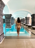 A woman in a swimsuit is walking towards a large indoor swimming pool situated in a grand architectural space with stone arches. The setting appears serene and luxurious, with the water reflecting the soft lighting from the arched ceiling.