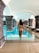 A woman in a swimsuit is walking towards a large indoor swimming pool situated in a grand architectural space with stone arches. The setting appears serene and luxurious, with the water reflecting the soft lighting from the arched ceiling.