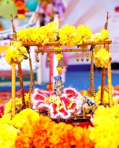 A decorative swing adorned with vibrant yellow marigold flowers holds a small, intricately dressed idol wearing a detailed headdress and colorful attire. In the background, there are blurred colors and patterns suggesting a festive or ceremonial setting.