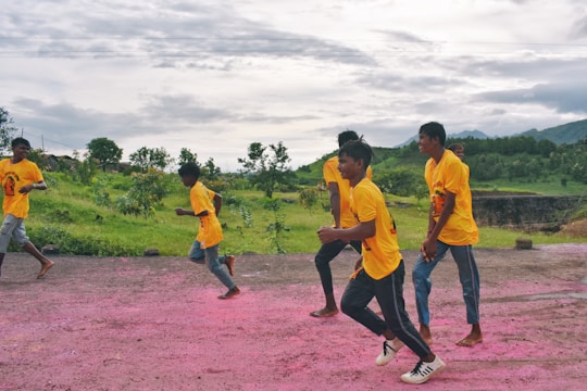 A group of young people wearing bright yellow shirts and jeans are running along a path with pink powder on the ground. The background features lush green scenery and cloudy skies, creating a vibrant and energetic scene.