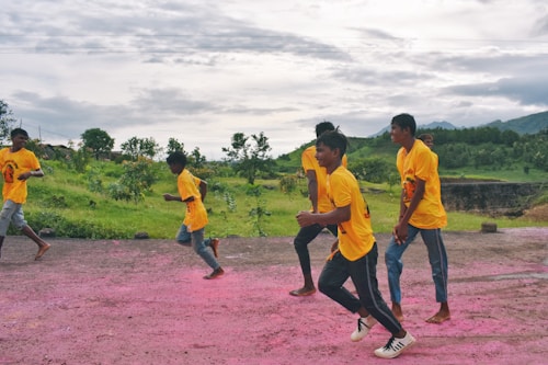 A group of young people wearing bright yellow shirts and jeans are running along a path with pink powder on the ground. The background features lush green scenery and cloudy skies, creating a vibrant and energetic scene.
