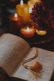 An open book with handwritten text lies on a table surrounded by several lit candles, creating a warm and intimate atmosphere. A gold-colored tassel serves as a bookmark, and flowers are visible in the background, adding a touch of elegance.