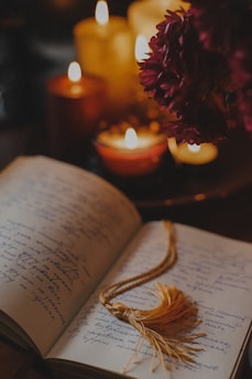 An open book with handwritten text lies on a table surrounded by several lit candles, creating a warm and intimate atmosphere. A gold-colored tassel serves as a bookmark, and flowers are visible in the background, adding a touch of elegance.
