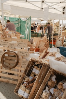 A bustling outdoor market scene featuring several stalls selling a variety of handmade and artisanal goods. Wooden crates and barrels are filled with items like pasta and baked goods. Several people are browsing and shopping under the shade of white tents, with an eclectic display of products. A backdrop of a striped green and white fabric adds color to the arrangement.