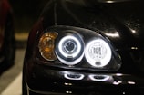 A close-up view of a car's headlight assembly at night. The headlights are illuminated, with circular LED lights and an amber turn signal visible. The car's hood is glossy and reflects light.
