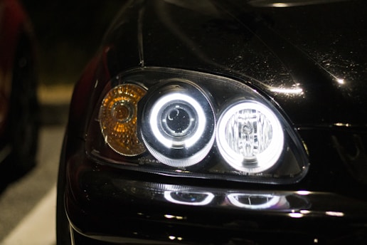 A close-up view of a car's headlight assembly at night. The headlights are illuminated, with circular LED lights and an amber turn signal visible. The car's hood is glossy and reflects light.