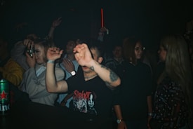 A group of people is gathered in a dimly lit environment, likely a nightclub or concert. The individuals are engaged in the event, with some raising their hands, while others are focused on capturing the moment with a smartphone. A person in a Slipknot t-shirt is prominently in the foreground, partially obscuring their face with a raised arm. The scene suggests an atmosphere of lively enjoyment and social interaction.