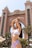 A woman in a bikini poses with her arms raised in front of a large, ornate building resembling a luxury hotel or resort. The background features palm trees, and the architecture is characterized by grand arches and intricate details.