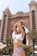 A woman in a bikini poses with her arms raised in front of a large, ornate building resembling a luxury hotel or resort. The background features palm trees, and the architecture is characterized by grand arches and intricate details.