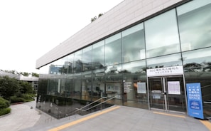 A modern building with large glass windows, reflecting the surrounding greenery and sky. The entrance has signage in Korean and English, indicating it is a makerspace. The building is surrounded by a well-maintained landscape with a pathway leading to the entrance.