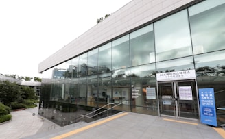 A modern building with large glass windows, reflecting the surrounding greenery and sky. The entrance has signage in Korean and English, indicating it is a makerspace. The building is surrounded by a well-maintained landscape with a pathway leading to the entrance.
