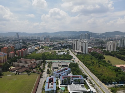 Aerial view of an urban landscape featuring a mix of residential and commercial high-rise buildings. Green areas, roads, and scattered trees are interspersed among the structures. The sky is partly cloudy, indicating a typical cityscape with distant hills in the background.