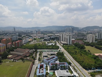 Aerial view of an urban landscape featuring a mix of residential and commercial high-rise buildings. Green areas, roads, and scattered trees are interspersed among the structures. The sky is partly cloudy, indicating a typical cityscape with distant hills in the background.