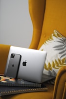 A sleek silver laptop and a black smartphone, both displaying a prominent Apple logo, are propped against the cushion of a mustard yellow armchair. The chair features a decorative cushion with gray and mustard leaf patterns. The devices rest on a couple of closed notebooks with a colorful design.