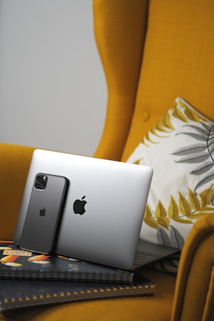 A sleek silver laptop and a black smartphone, both displaying a prominent Apple logo, are propped against the cushion of a mustard yellow armchair. The chair features a decorative cushion with gray and mustard leaf patterns. The devices rest on a couple of closed notebooks with a colorful design.