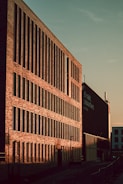 A large brick building with numerous vertical windows is bathed in warm sunlight, casting strong shadows. The sign on the building indicates it is a market. The sky is clear and the street is empty.
