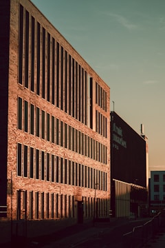 A large brick building with numerous vertical windows is bathed in warm sunlight, casting strong shadows. The sign on the building indicates it is a market. The sky is clear and the street is empty.