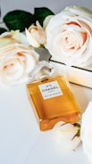 A rectangular glass perfume bottle with a golden amber color liquid inside is placed on a white surface. The label on the bottle reads 'N°5 Chanel Paris Eau de Parfum'. Surrounding the bottle are several blooming ivory roses, adding a touch of elegance and luxury.