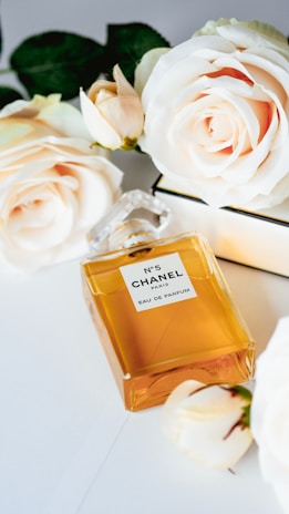 A rectangular glass perfume bottle with a golden amber color liquid inside is placed on a white surface. The label on the bottle reads 'N°5 Chanel Paris Eau de Parfum'. Surrounding the bottle are several blooming ivory roses, adding a touch of elegance and luxury.