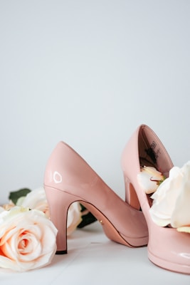 A pair of elegant pink high-heeled shoes is surrounded by soft pastel roses. The shoes have a glossy finish, creating a reflective shine that complements the delicate petals of the roses.