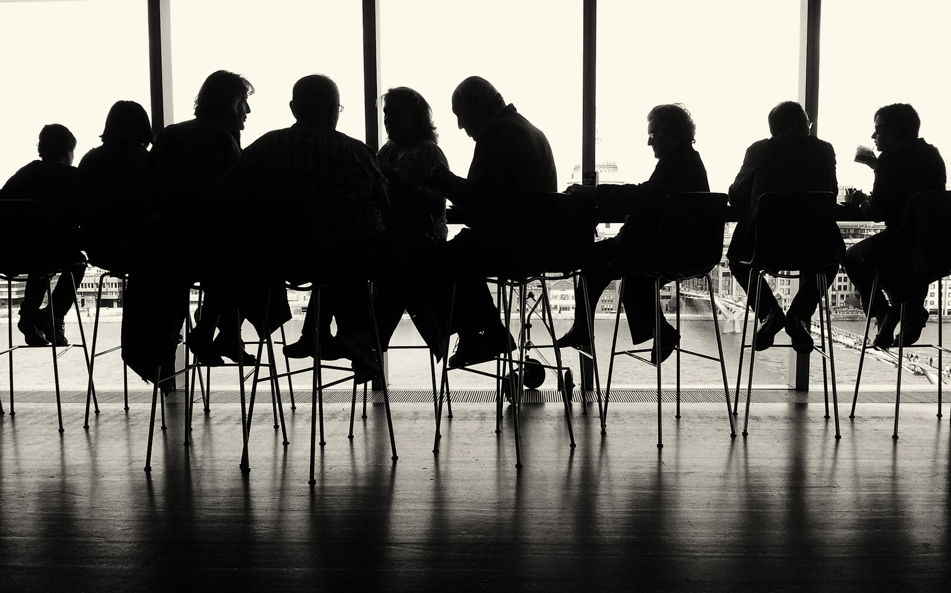 Equipo reunido alrededor de una mesa alta, visto a contraluz frente a un ventanal, en blanco y negro.