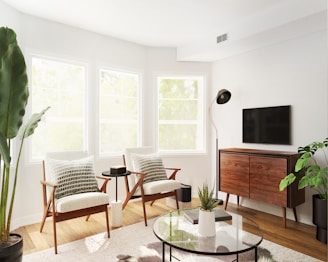 A modern living room with two mid-century modern chairs placed near three large windows allowing natural light to flood in. The room features a wooden console with a TV mounted above it. A sleek floor lamp stands next to the TV. The decor includes indoor plants and a glass coffee table with a round, black base. The overall theme is minimalistic with a mix of natural and contemporary elements.