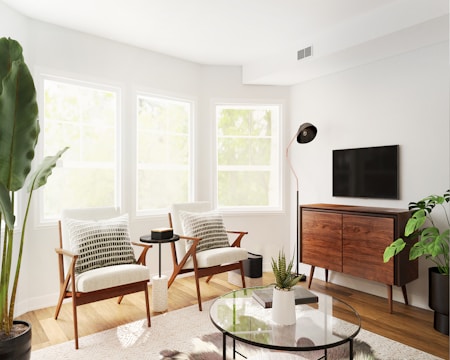 A modern living room with two mid-century modern chairs placed near three large windows allowing natural light to flood in. The room features a wooden console with a TV mounted above it. A sleek floor lamp stands next to the TV. The decor includes indoor plants and a glass coffee table with a round, black base. The overall theme is minimalistic with a mix of natural and contemporary elements.