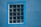 A window composed of 20 glass blocks is set into a blue stucco wall. The window frame is a lighter shade of blue, providing a contrast against the wall. The texture of the wall is rough, while the glass blocks have a clear, reflective surface that slightly distorts the view through them.