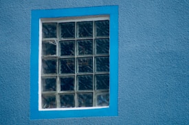 A window composed of 20 glass blocks is set into a blue stucco wall. The window frame is a lighter shade of blue, providing a contrast against the wall. The texture of the wall is rough, while the glass blocks have a clear, reflective surface that slightly distorts the view through them.
