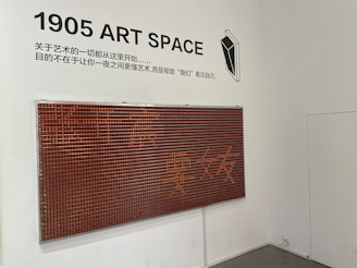 A white wall featuring a modern art installation with a grid of small red and black squares forming a large rectangular panel. Above it, black text reads '1905 ART SPACE' alongside Chinese characters. A minimalistic black and white geometric illustration is also present.