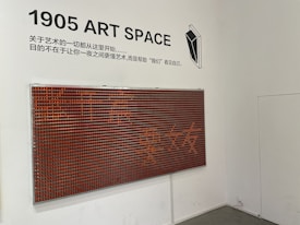 A white wall featuring a modern art installation with a grid of small red and black squares forming a large rectangular panel. Above it, black text reads '1905 ART SPACE' alongside Chinese characters. A minimalistic black and white geometric illustration is also present.