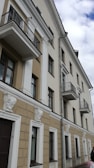 A multi-story residential building with a beige and white facade featuring classical architecture elements such as decorative moldings and small balconies with metal railings. The walls are adorned with ornamental sculptures, and there are several rectangular windows visible.