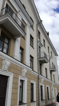 A multi-story residential building with a beige and white facade featuring classical architecture elements such as decorative moldings and small balconies with metal railings. The walls are adorned with ornamental sculptures, and there are several rectangular windows visible.