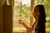 A person with shoulder-length hair is smiling while placing an envelope into a mailbox inside a building. Sunlight streams in from a window, casting a warm, golden glow throughout the scene. The background reveals a green, blurred view outside the window.