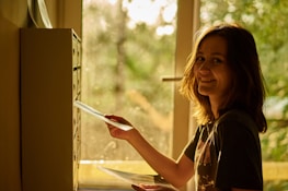 A person with shoulder-length hair is smiling while placing an envelope into a mailbox inside a building. Sunlight streams in from a window, casting a warm, golden glow throughout the scene. The background reveals a green, blurred view outside the window.