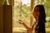 A person with shoulder-length hair is smiling while placing an envelope into a mailbox inside a building. Sunlight streams in from a window, casting a warm, golden glow throughout the scene. The background reveals a green, blurred view outside the window.