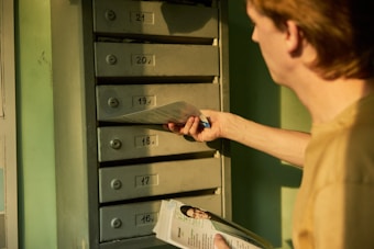 A person is holding and inserting an envelope into a vertical arrangement of mailboxes numbered from 16 to 21. The mailboxes appear metallic and are located against a green-hued wall. The person's focus is on the task, holding another item, possibly a magazine, in their other hand.