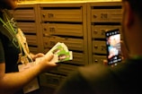 A person holding several flyers with images of a man in front of a wall of numbered mailboxes. Another person is taking a photo with a smartphone.
