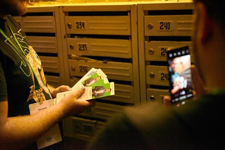 A person holding several flyers with images of a man in front of a wall of numbered mailboxes. Another person is taking a photo with a smartphone.