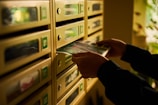 A person is placing or retrieving mail from a row of metallic mailboxes. The boxes are numbered, and there are small windows on each. The lighting is warm, suggesting a cozy or dimly lit environment.