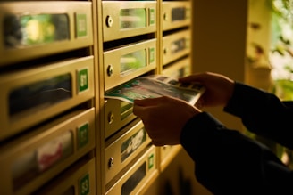 A person is placing or retrieving mail from a row of metallic mailboxes. The boxes are numbered, and there are small windows on each. The lighting is warm, suggesting a cozy or dimly lit environment.