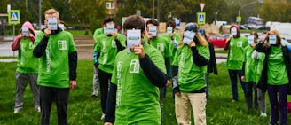 A group of people stand on a grassy area, all wearing green T-shirts with white text or logos. Each person is holding up a badge or sign that obscures their face. The background includes a road, parked cars, and some trees, indicating an outdoor setting.