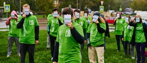 A group of people stand on a grassy area, all wearing green T-shirts with white text or logos. Each person is holding up a badge or sign that obscures their face. The background includes a road, parked cars, and some trees, indicating an outdoor setting.