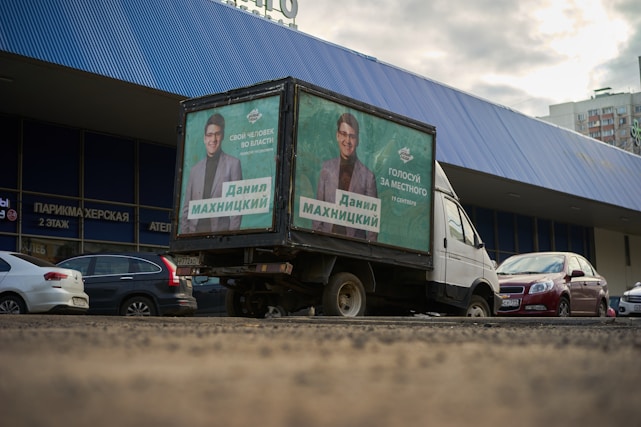A truck with political campaign posters is parked in front of a building with a blue corrugated metal facade. Several cars are also parked nearby. The posters depict a man in a suit, and the text suggests support for a local candidate. The scene appears to be set in an urban area, with apartment buildings visible in the background and a slightly overcast sky.
