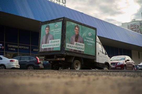 A truck with political campaign posters is parked in front of a building with a blue corrugated metal facade. Several cars are also parked nearby. The posters depict a man in a suit, and the text suggests support for a local candidate. The scene appears to be set in an urban area, with apartment buildings visible in the background and a slightly overcast sky.