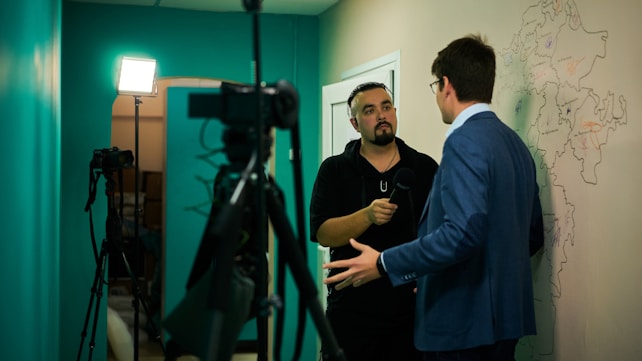 Two people are engaged in a conversation or interview in a room with teal-colored walls. One person, holding a microphone, is wearing a black hoodie. The other, dressed in a blue suit, stands against a wall with a map drawn on it. There are cameras and lighting equipment set up, suggesting a professional or journalistic setup.